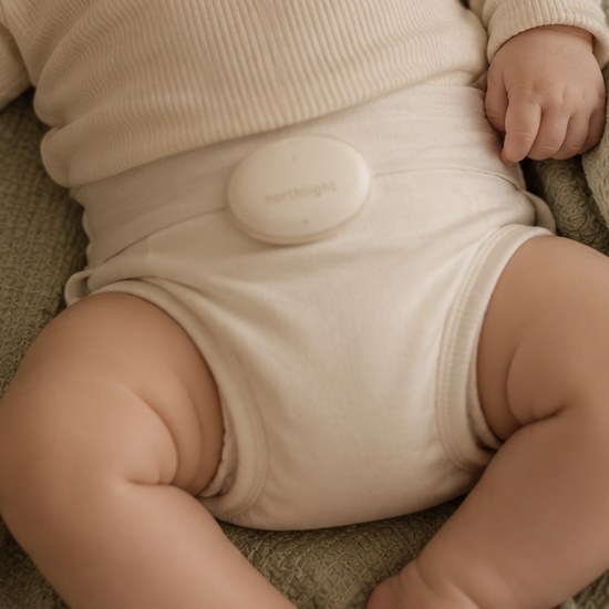 The Northlight sensor, a small soft bone-white pebble embossed with the northlight mark, clipped gently to a sleeping baby's diaper waistband.